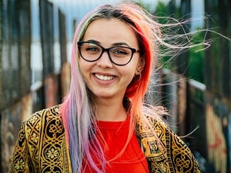 Tjej med röd topp och regnbågsfärgat hår står vid en husvägg