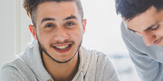 En mörkhårig kille tittar in i kameran, en annan skymtar fram