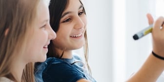 Två flickor samarbetar vid en whiteboard. De ser engagerade ut och arbetar tillsammans med en uppgift
