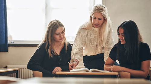 En lärare står mellan två elever vid en rad skolbänkar