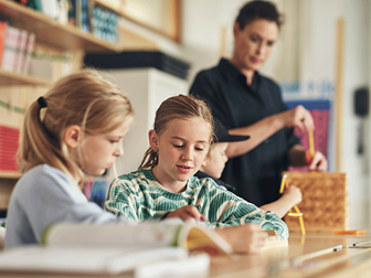 Två mellanstadietjejer pluggar vid sina bänkar. En lärare i bakgrunden
