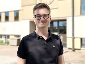 Ung man med glasögon i svart tröja, står utomhus och ler mot kameran